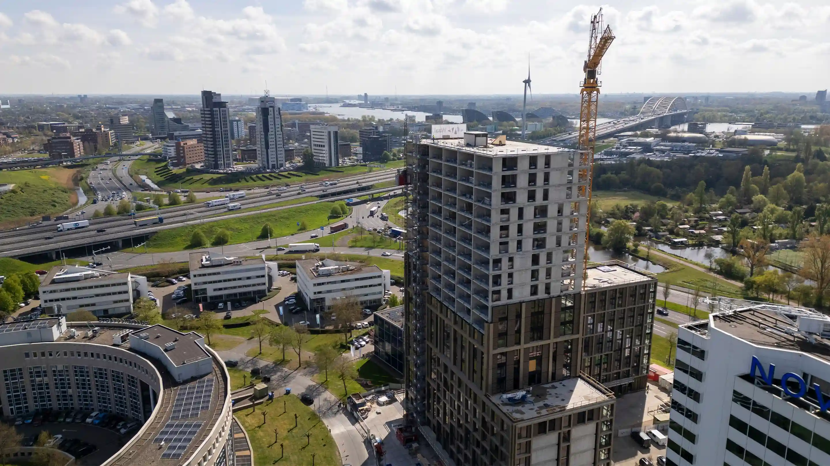 Aerial view of the high-rise residential project Canvas Living under construction at Brainpark Rotterdam, with a crane on top of the building and the Van Brienenoord Bridge and surrounding cityscape in the background.