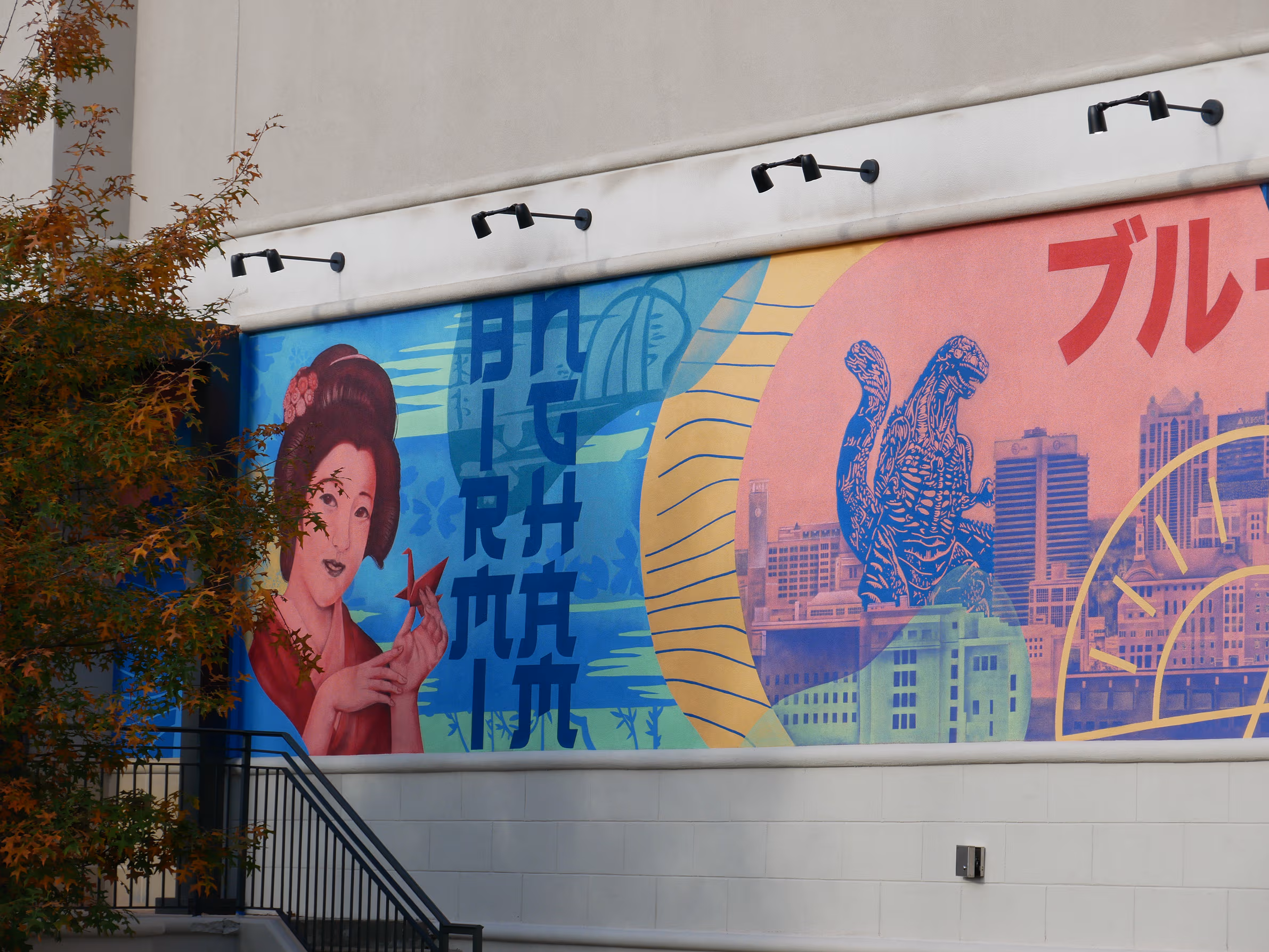 A wide shot of a mural painted on a side of a sushi shop building featuring vibrant blue, yellow and red colors including Japanese themed elements like a Japanese geisha and Godzilla.