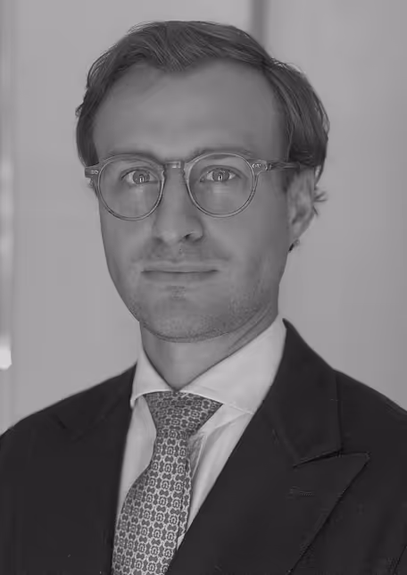 Portrait of a man wearing round glasses, a dark suit, white shirt, and patterned tie, looking at the camera.
