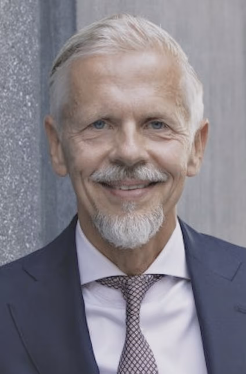 Smiling elderly man with white hair and beard wearing a dark suit, white shirt, and patterned tie against a gray background.