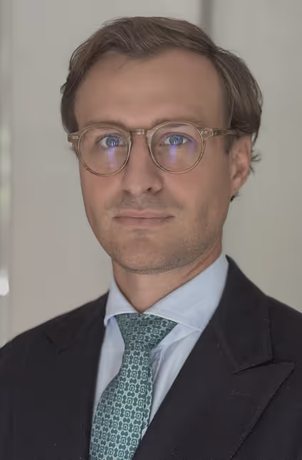 Man with light brown hair and round glasses wearing a dark suit jacket, white shirt, and green patterned tie.