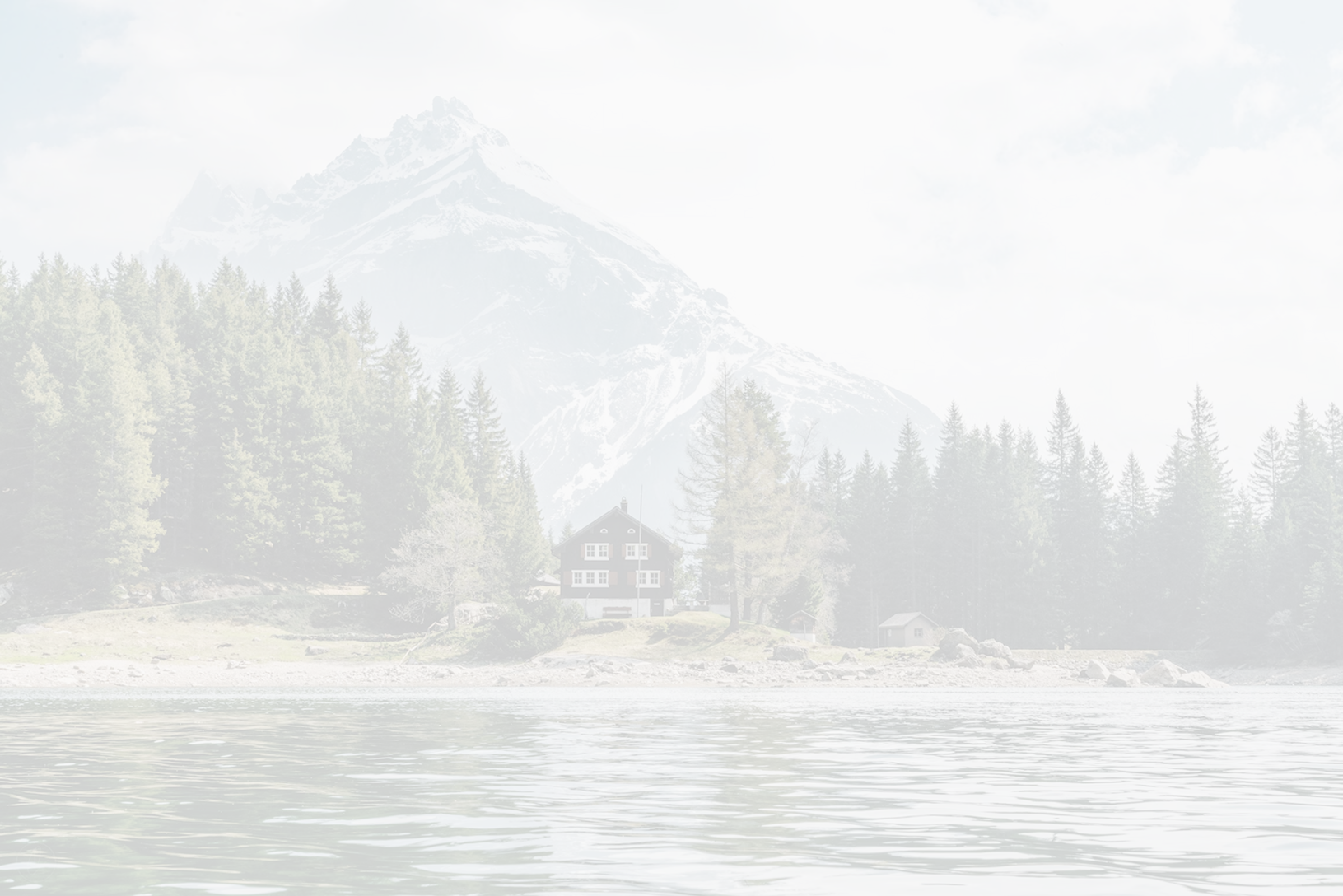 Lake with rippling water in the foreground, dense evergreen trees, a small cabin, and snow-capped mountain in the background under a cloudy sky.