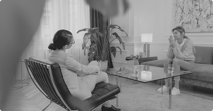 Two women engaged in a conversation in a modern living room, one sitting on a leather chair and the other on a sofa across a glass coffee table.
