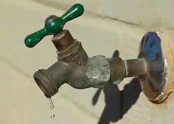 A close-up, low-angle shot captures an old, weathered outdoor spigot with a green handle, leaking water in distinct drops. The spigot is mounted on a light-colored wall, and the surrounding area shows signs of wear and tear, including chipped paint and ru