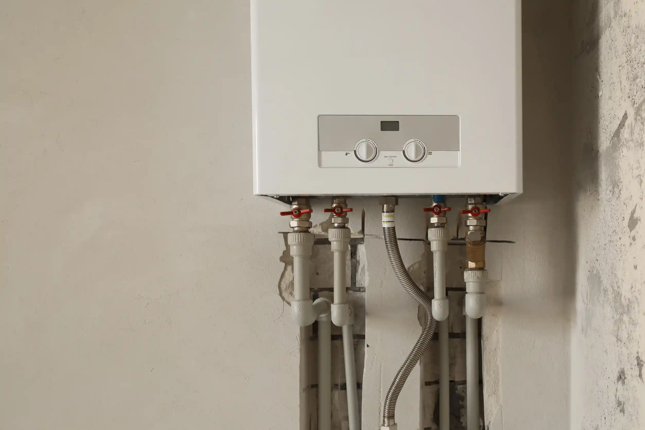 A white tankless water heater is mounted on a light grey wall, with multiple pipes and red and blue valves extending from its bottom. The wall surrounding the pipes appears to be in a state of renovation or unfinished construction.