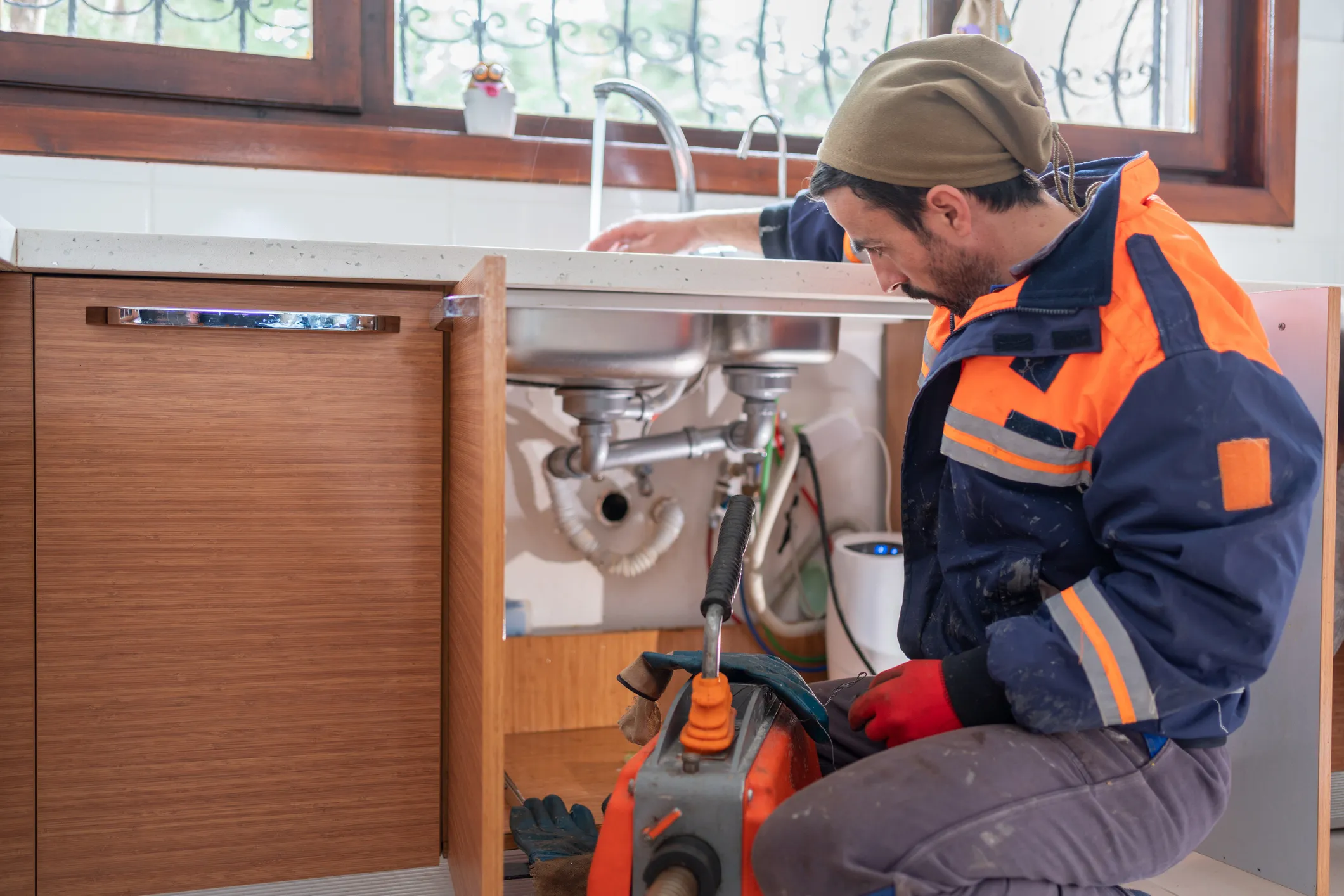 A plumber wearing a jacket is repairing a sink using tools
