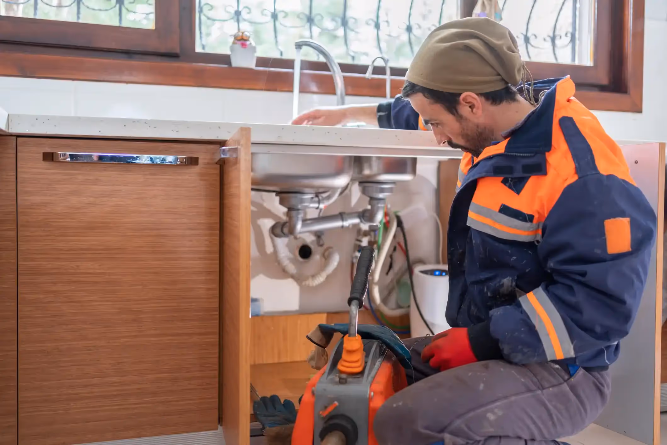 A male plumber wearing work clothes and a cap is kneeling and working on the pipes under a kitchen sink.