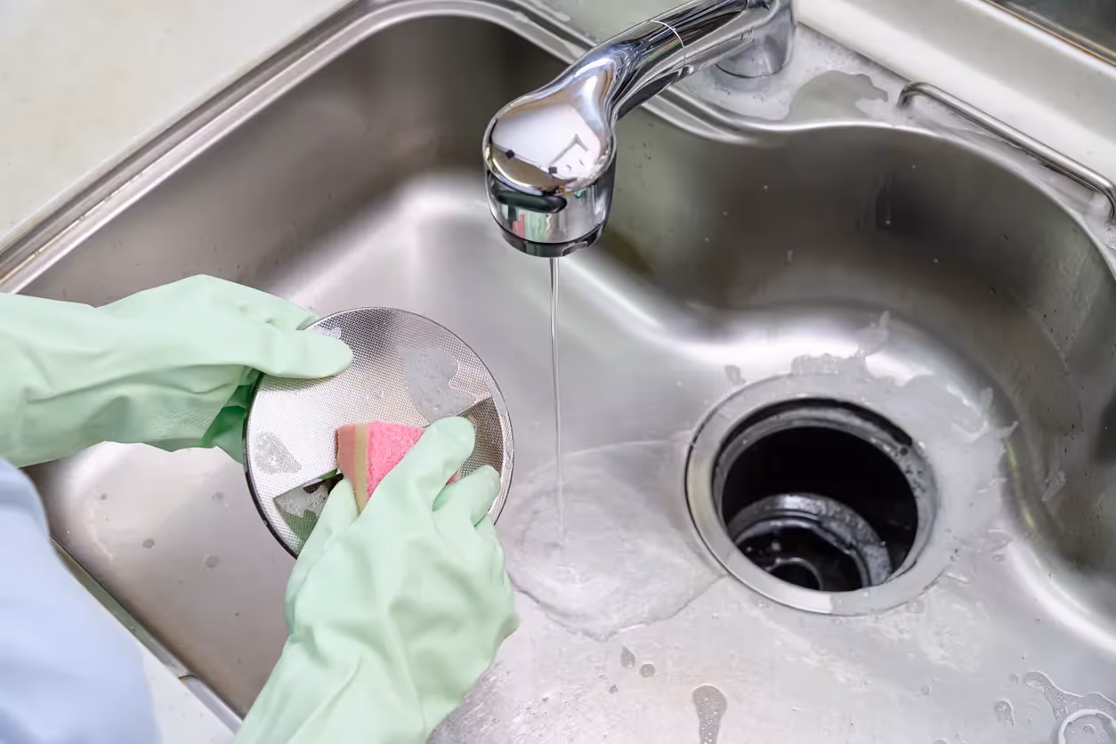 A person wearing light green rubber gloves is cleaning a metal sink stopper with a pink sponge under running water in a stainless steel kitchen sink.