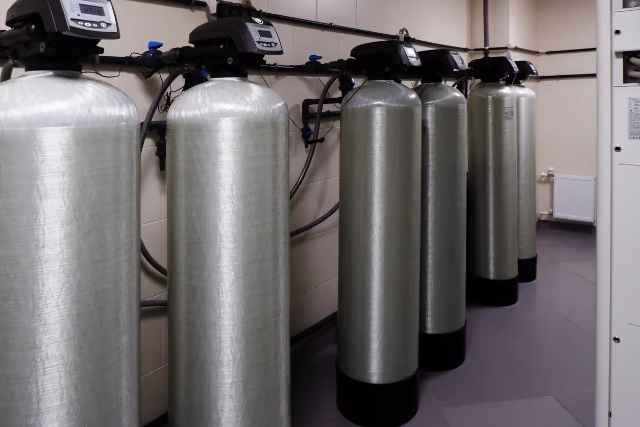 A row of large, cylindrical water softening or filtration tanks with digital controls on top, connected by black pipes, in what appears to be an industrial or commercial setting.