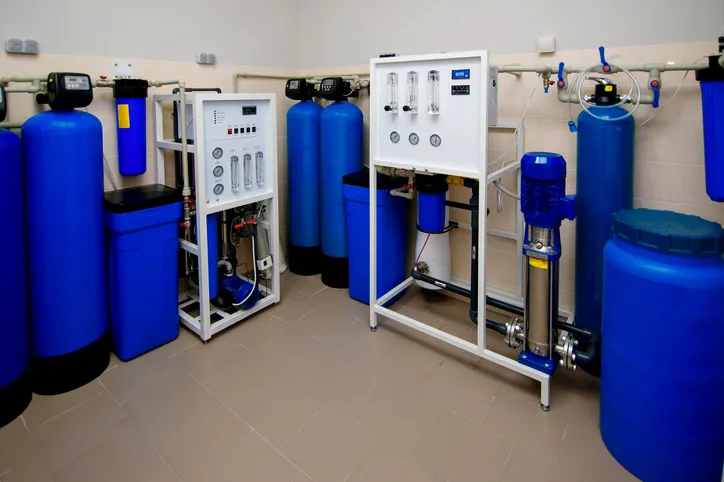 A comprehensive industrial or commercial water treatment room featuring multiple large blue filter tanks, control panels, and pumping equipment.