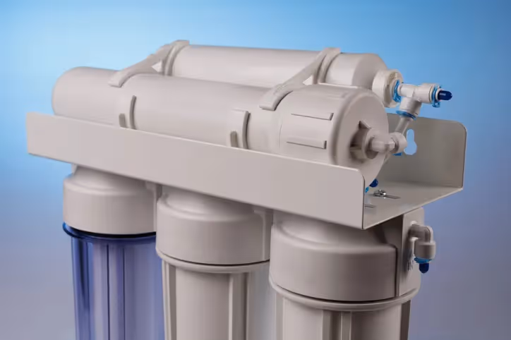  A close-up studio shot of a reverse osmosis water filter system. The unit is composed of four white cylindrical filter cartridges and one transparent blue cartridge, all mounted on a white metal frame. 