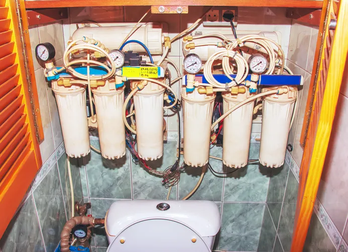 A multi-stage water filtration system with six vertical filters and prominent pressure gauges, mounted on a tiled wall above a toilet in a utility space.