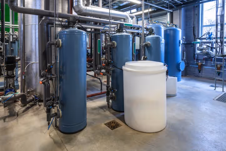 A large-scale industrial water treatment facility featuring multiple tall, blue and silver filter tanks, complex piping, and a large white cylindrical tank on a concrete floor.