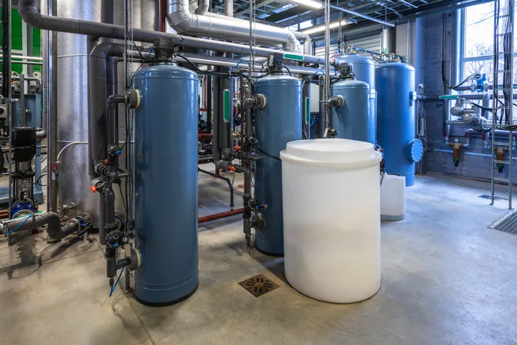 A large-scale industrial water treatment facility featuring multiple tall, blue and silver filter tanks, complex piping, and a large white cylindrical tank on a concrete floor.