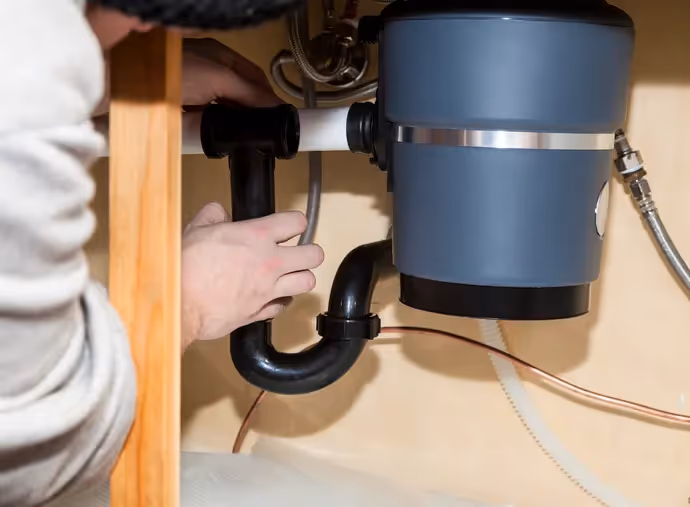 Close-up of a person connecting the black P-trap pipe to a gray garbage disposal unit under a kitchen sink.