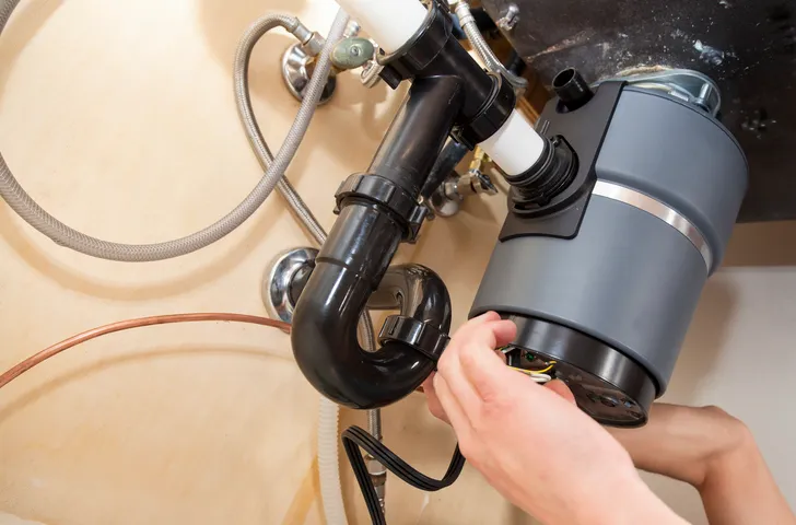 Person connecting electrical wires to the bottom of a gray garbage disposal unit under a sink with P-trap visible.
