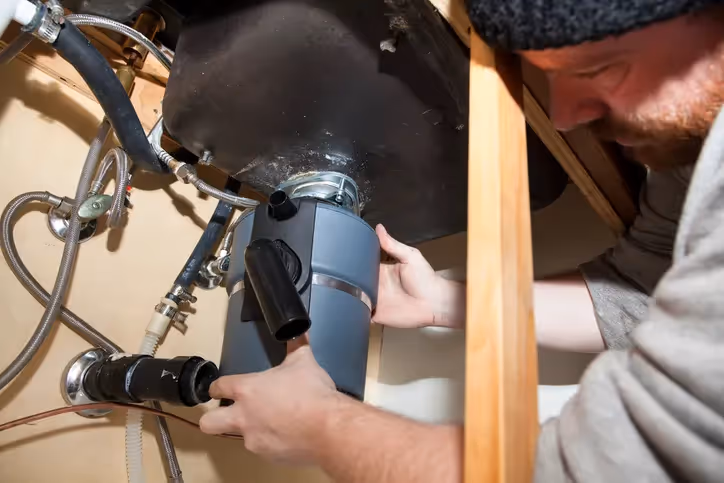 Man with a beard installing a gray garbage disposal under a sink, connecting the drain pipe.