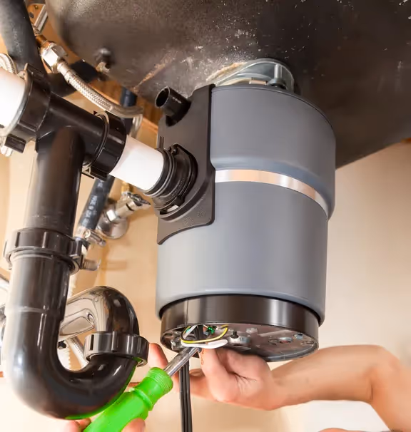 Hands wiring the electrical connections of a new gray garbage disposal unit under a kitchen sink with a green screwdriver.