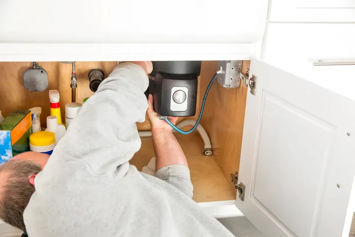 Man reaching into a kitchen cabinet to install or work on a black garbage disposal unit connected to the sink.