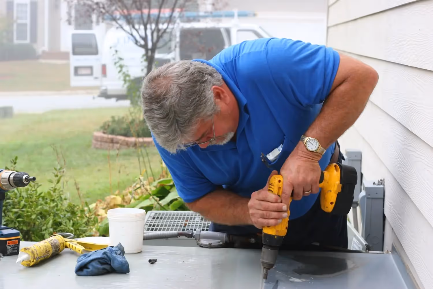 A man in a blue shirt and glasses uses a power drill on an air conditioning unit outdoors.
