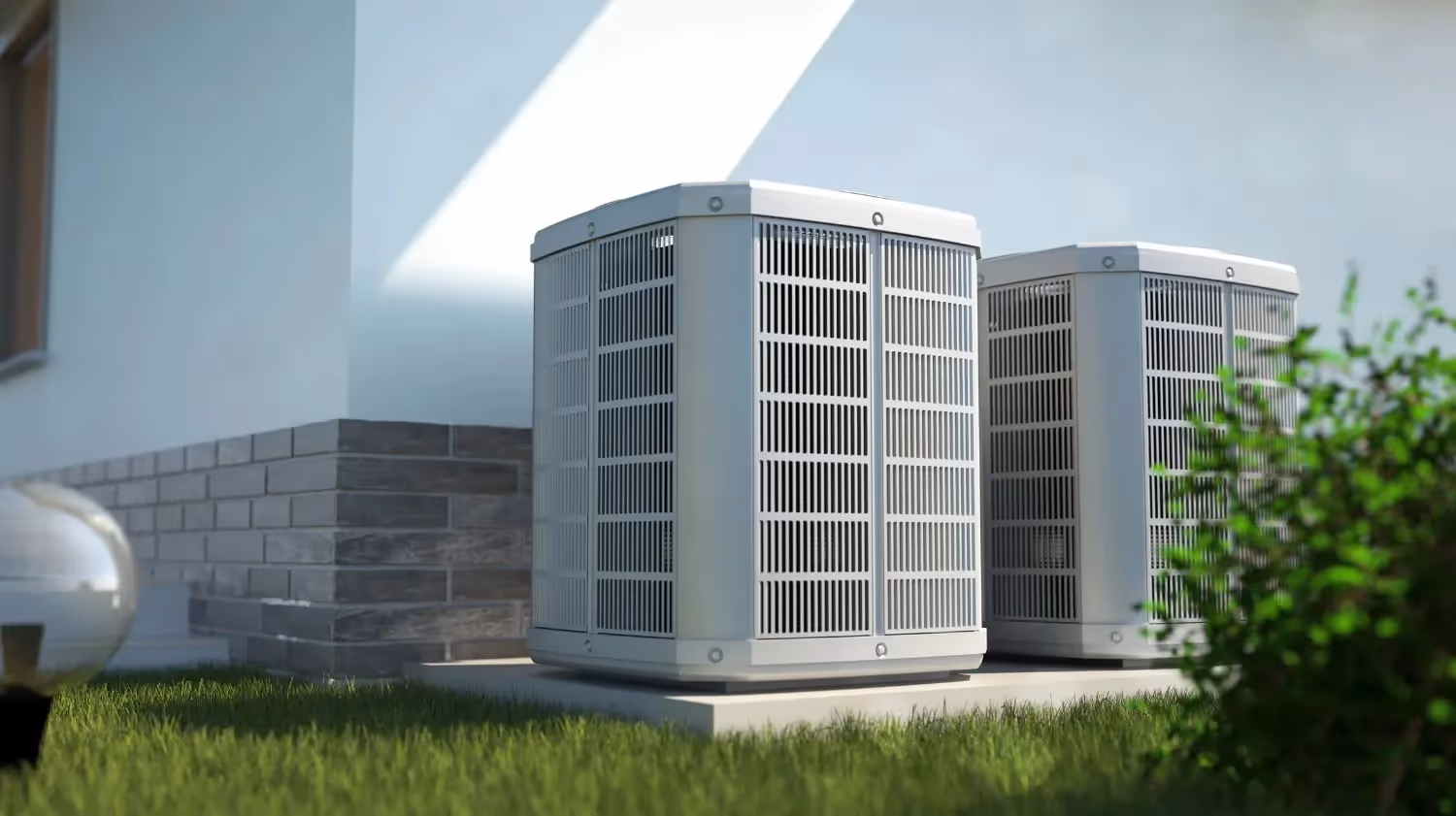Two outdoor air conditioning or heat pump units sit on a concrete pad next to a house with green bushes and grass.