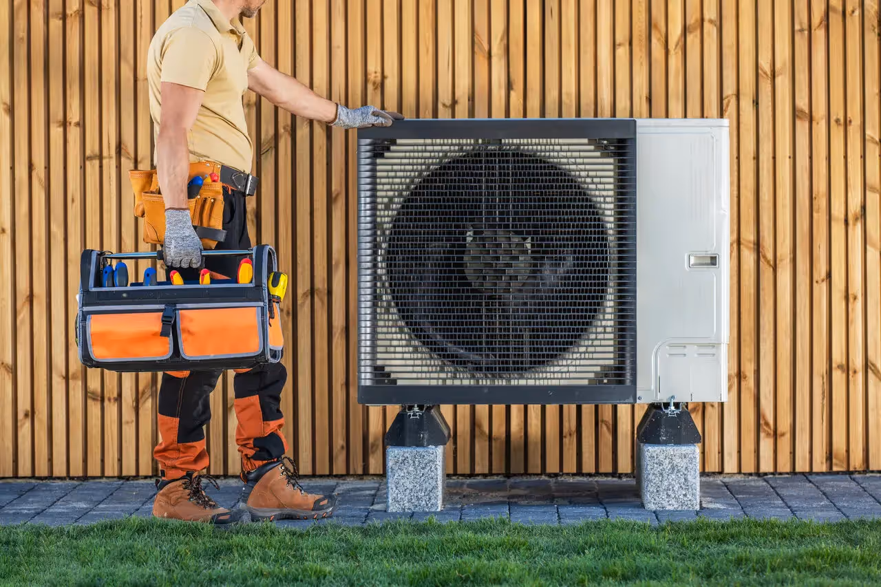 A technician stands next to a large heat pump, holding a toolbox, with a wooden fence in the background.