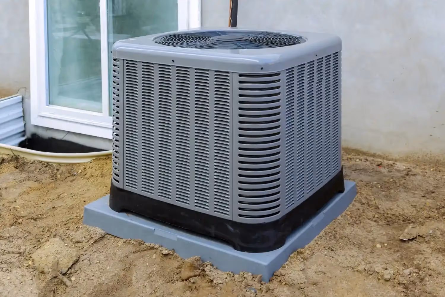A gray outdoor air conditioning unit sits on a plastic pad on a dirt and gravel surface next to a house.