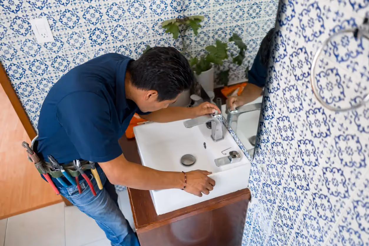 A plumber in a blue polo shirt and jeans with tools on his belt leans over a white bathroom sink, working on the faucet.
