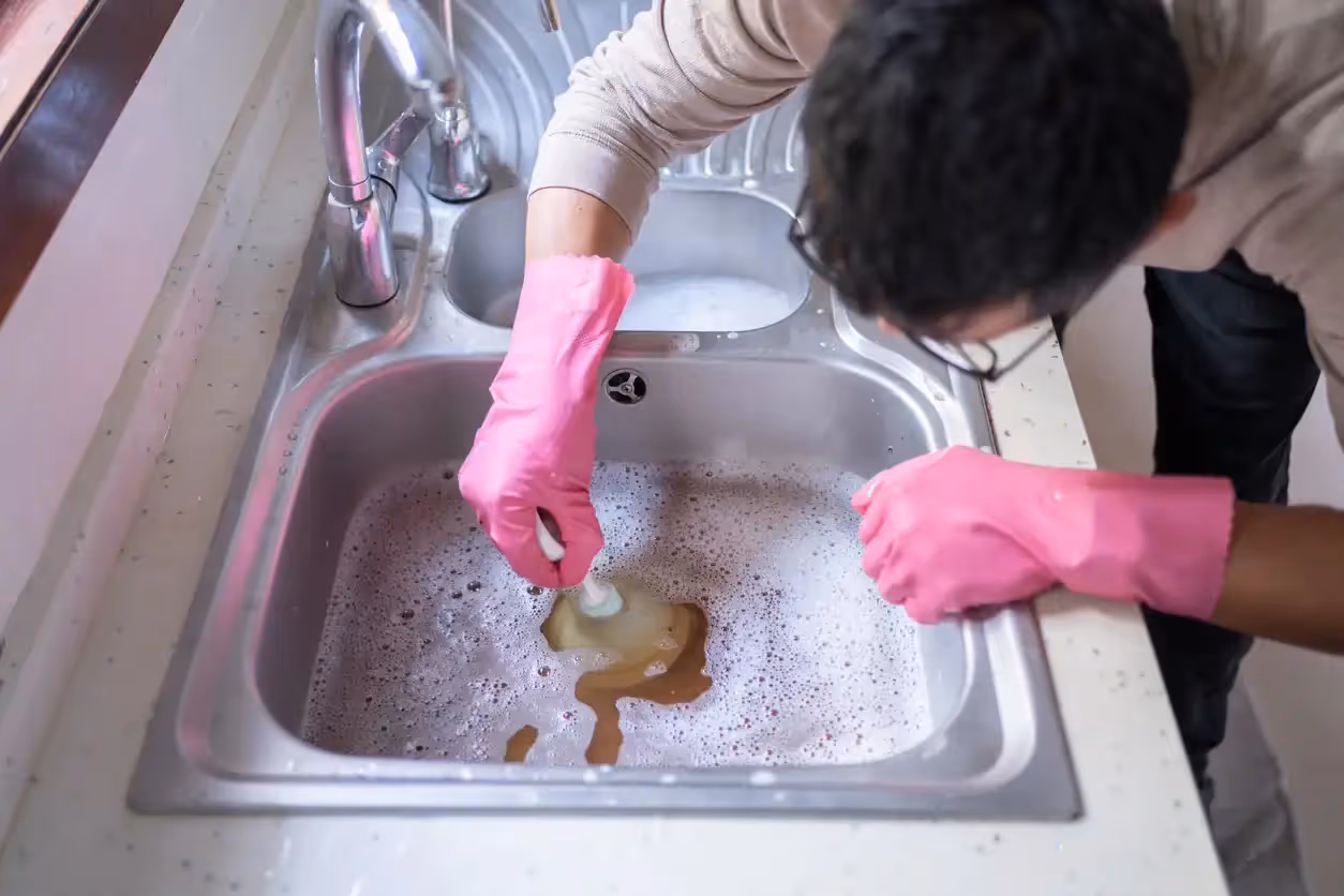 A person wearing pink gloves cleans a stainless steel kitchen sink filled with soapy, brown water.