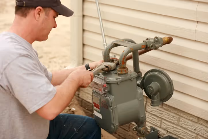 A man in a grey t-shirt and baseball cap uses a wrench to work on a grey gas meter and its associated piping, located outdoors against a beige wall.