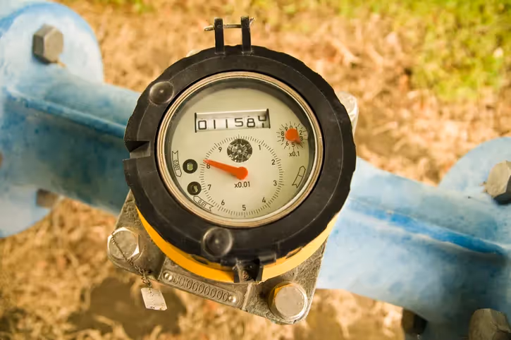 A close-up, overhead view of a black-cased water meter with a white face and digital readout, attached to a large blue pipe on a dirt and grass background.
