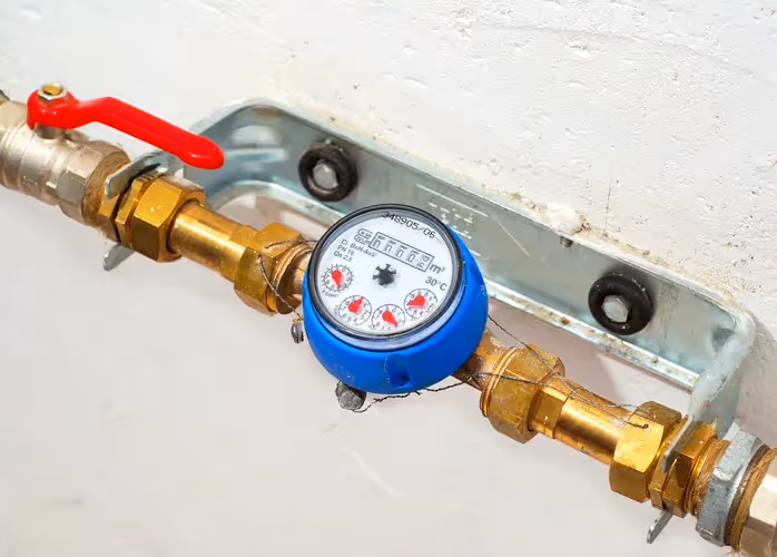 A close-up of a blue-cased water meter attached to brass pipes, featuring a red valve handle and mounted on a white wall.