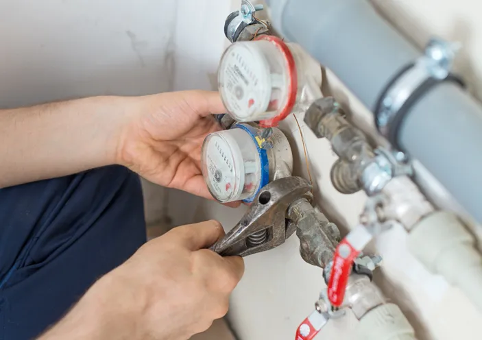 A person's hands work on adjusting a water meter using a wrench, with another meter and grey and white pipes visible in the background.