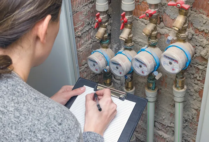 A person from behind is seen writing on a clipboard, taking readings from a row of four water meters with brass fittings and red valves on a brick wall.