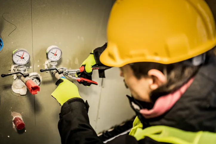 A person in a yellow hard hat and neon yellow gloves uses pliers to work on a water pipe connection next to two pressure gauges.