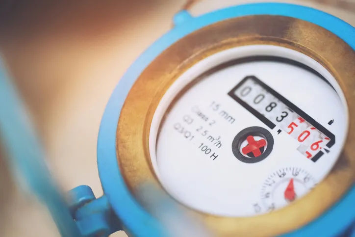A close-up, slightly blurred image of a blue-cased water meter's display, showing numbers and a small red cross, with brass tones in the background.