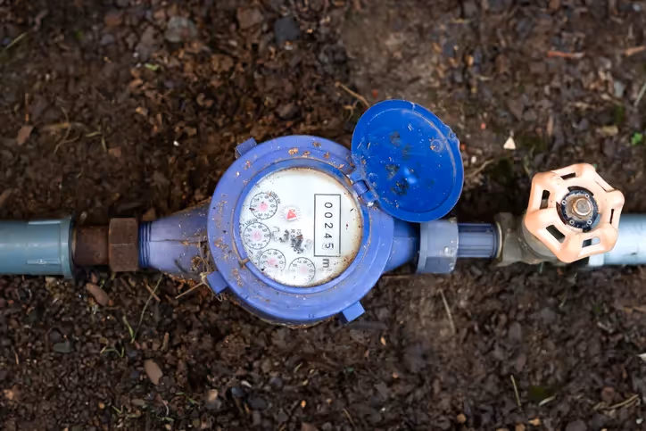 A person's hands work on adjusting a water meter using a wrench, with another meter and grey and white pipes visible in the background.