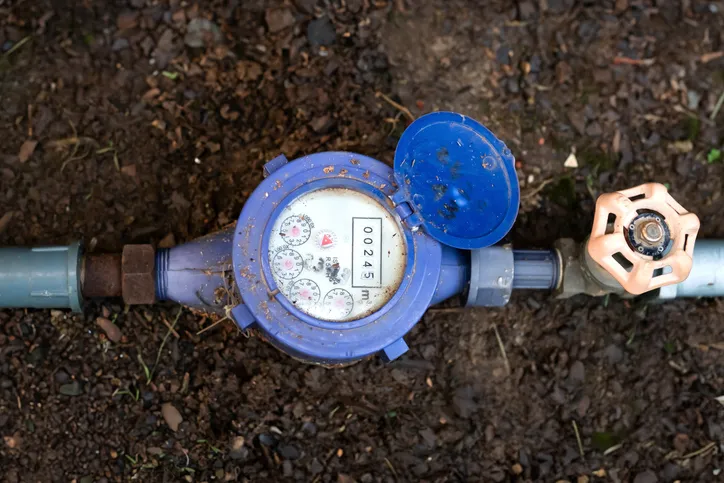 An overhead view of a blue water meter with its lid open, showing a digital readout, connected to a pipe system in the dirt with a faded orange valve.
