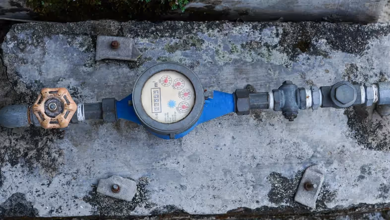 An overhead view of a blue water meter and an orange valve connected by metal pipes, mounted on a weathered concrete surface with rusty bolts.