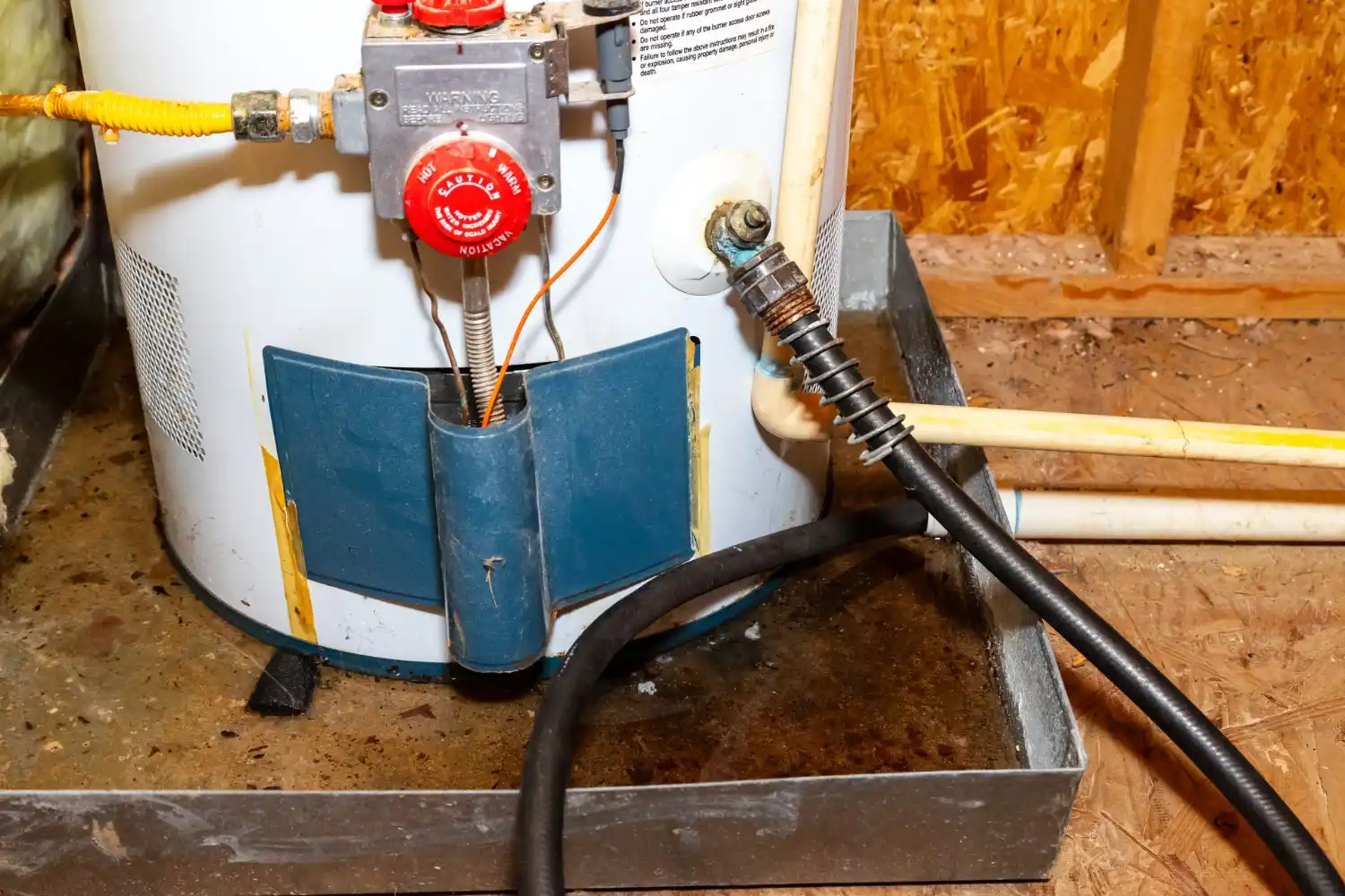 A close-up of the bottom of a white hot water heater shows a black hose, rusted pipes, and a metal basin collecting leaked water on a wooden floor.