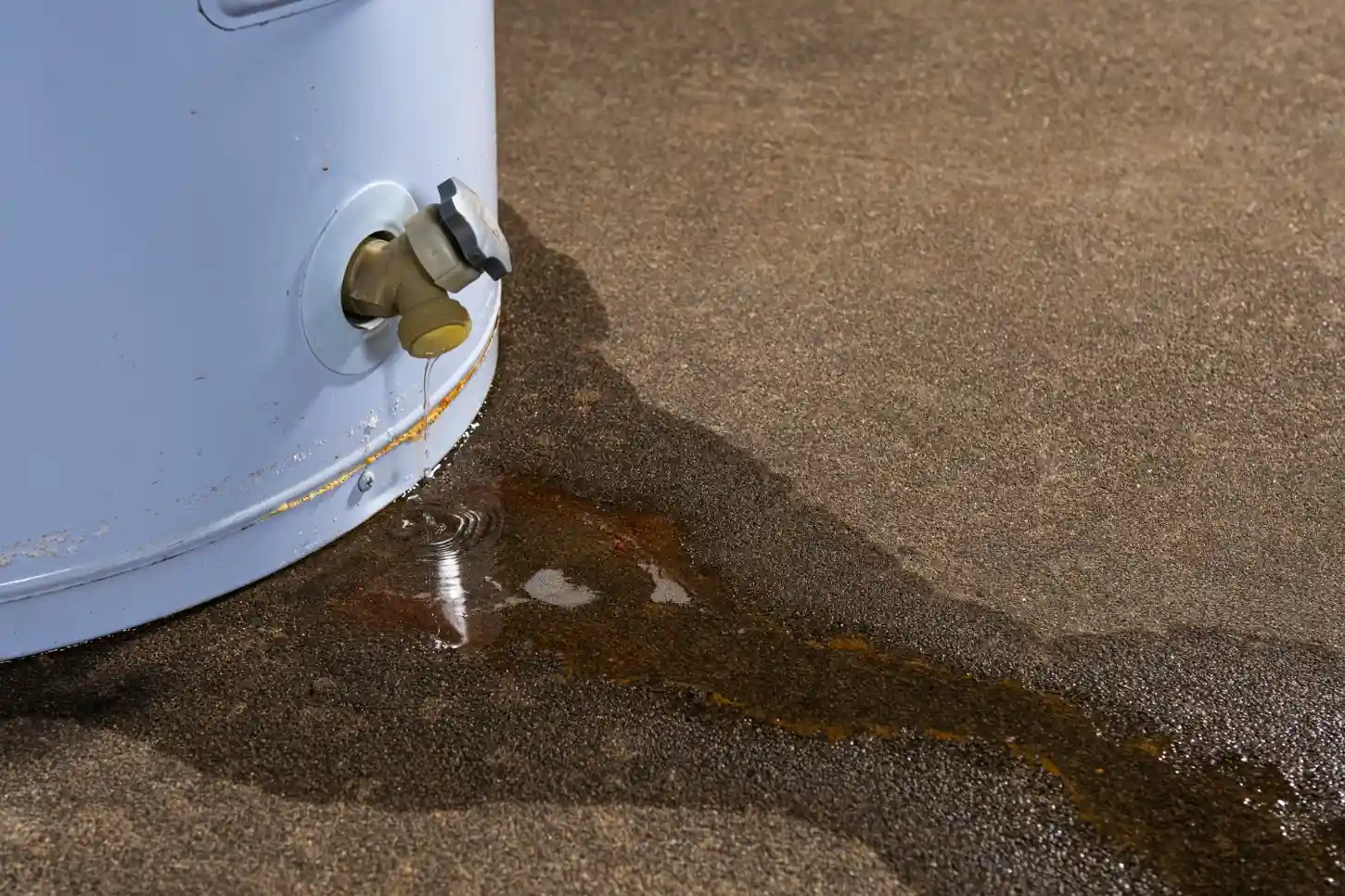 A close-up of the bottom of a white hot water heater shows water dripping from a yellow valve and forming a rusty-looking puddle on the concrete floor.
