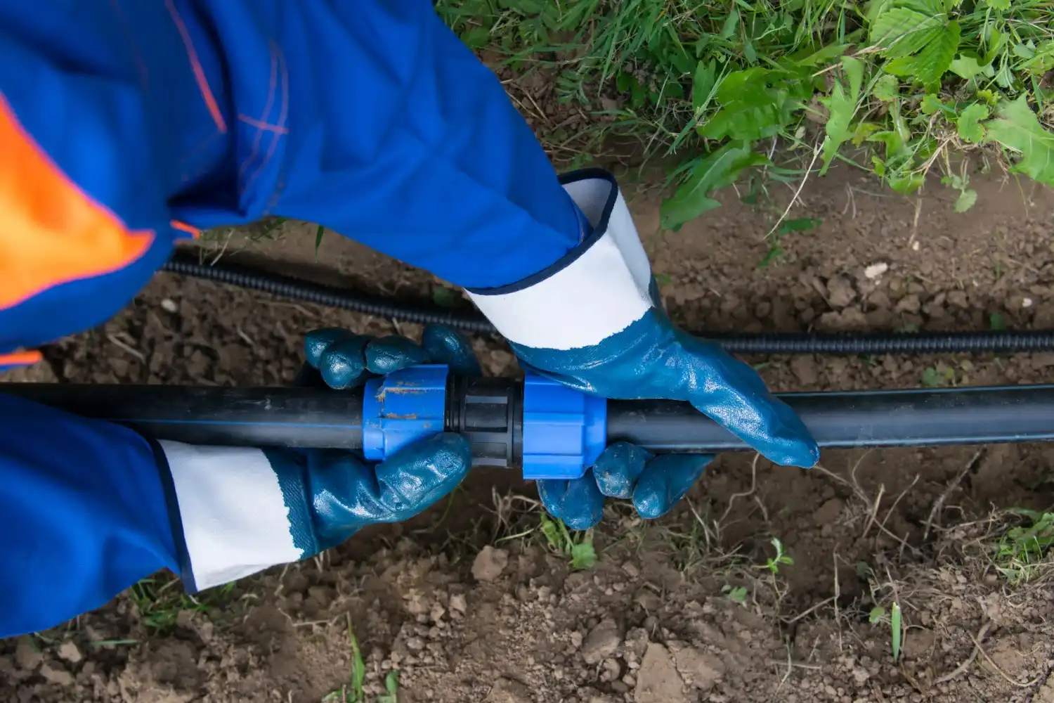 A worker in a blue uniform and gloves connects a blue compression fitting to a black water pipe laid in a shallow trench.