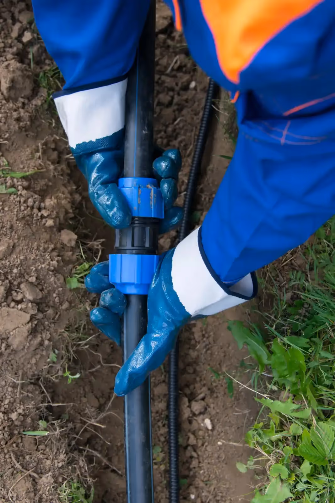 A close-up shot of a worker's gloved hands connecting a black plastic water pipe with a blue compression fitting in a trench.