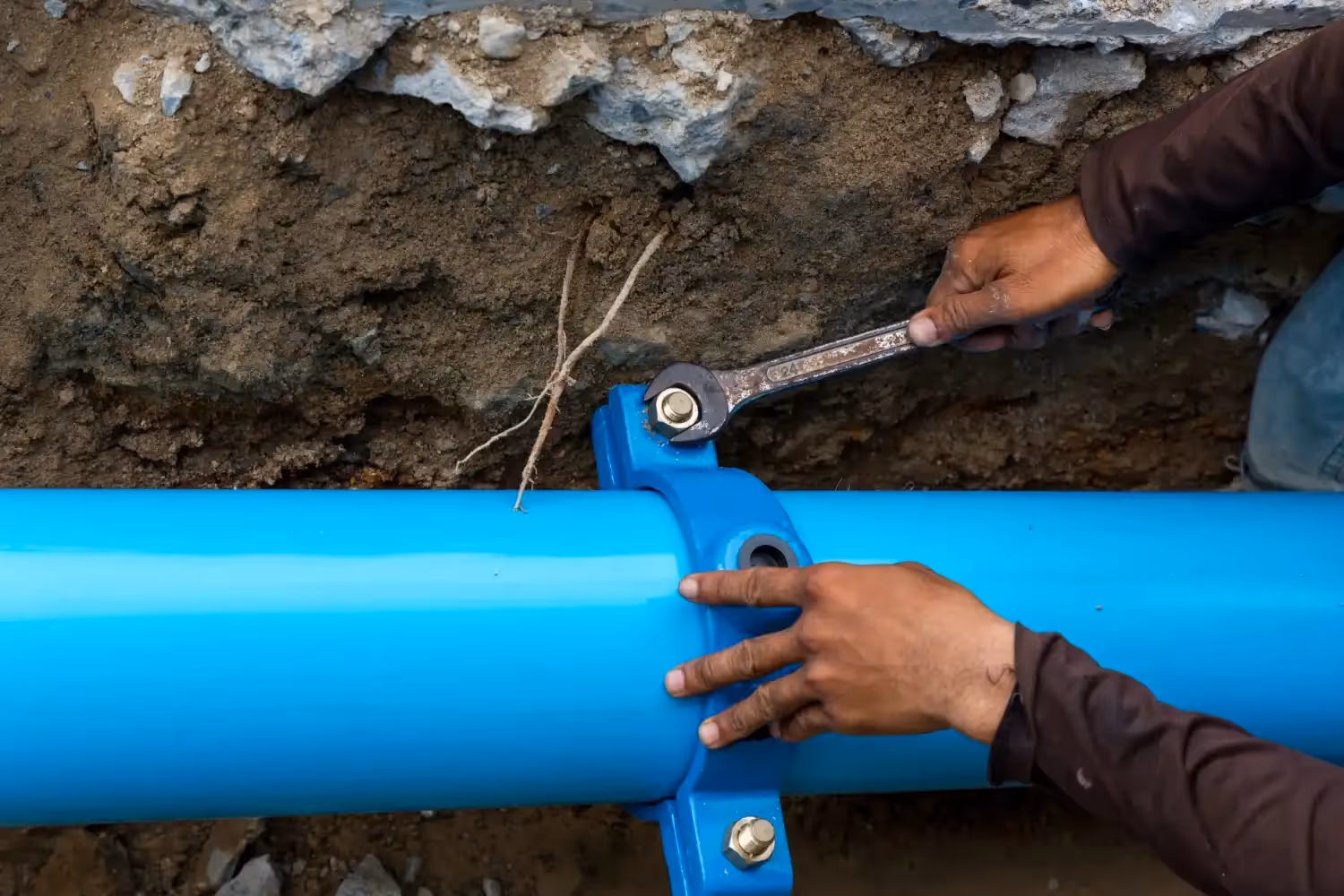 A worker's hands use a wrench to tighten a bolt on a blue pipe clamp, securing a large blue water line in a ditch.