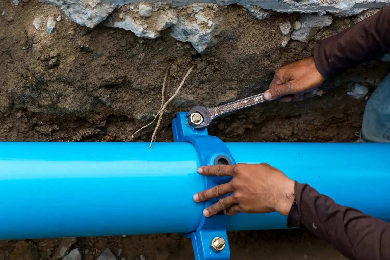 A worker's hands use a wrench to tighten a bolt on a blue pipe clamp, securing a large blue water line in a ditch.