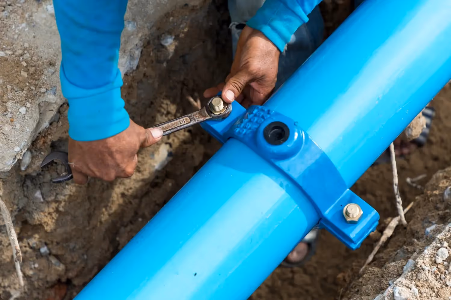A close-up of a worker's hands using a wrench to tighten a bolt on a blue saddle clamp attached to a large blue water pipe.