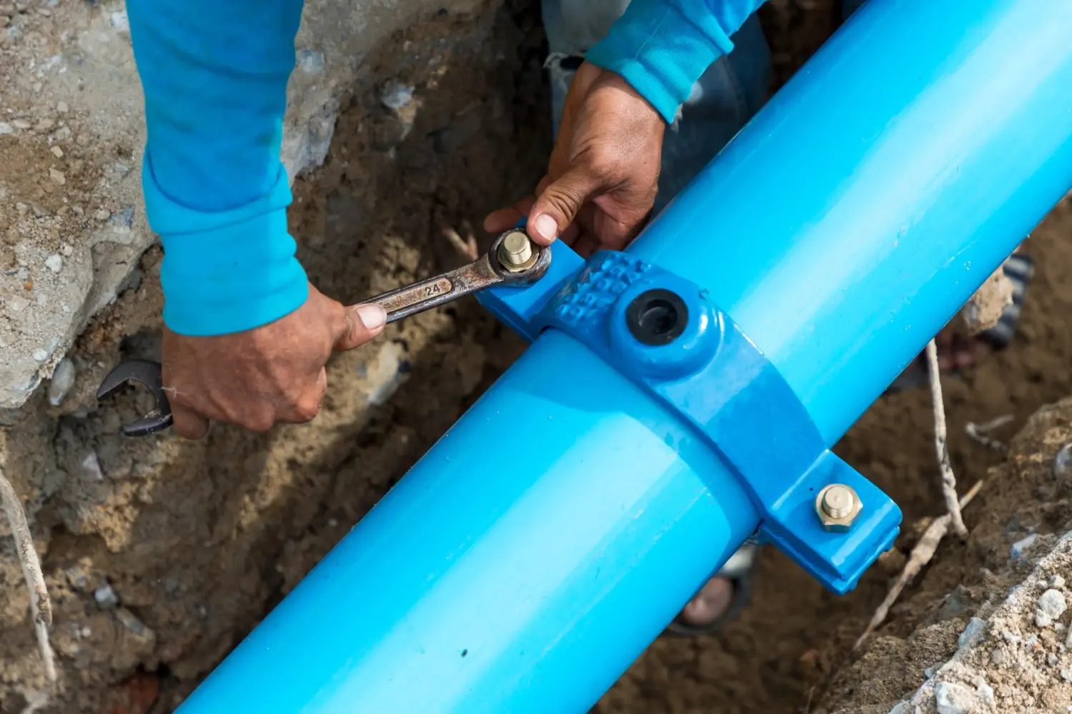 A close-up of a worker's hands using a wrench to tighten a bolt on a blue saddle clamp attached to a large blue water pipe.