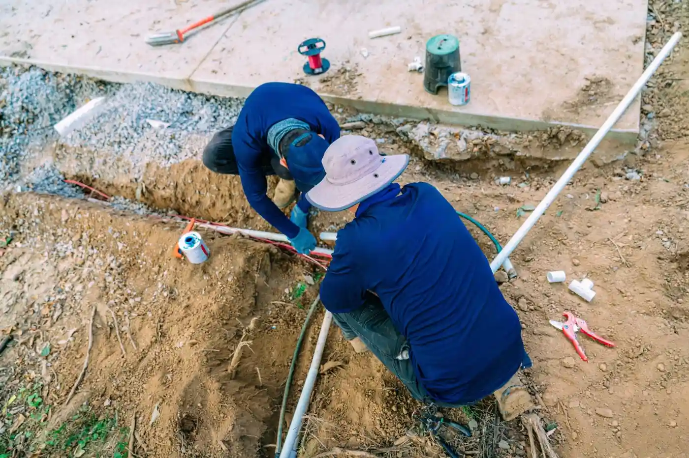 Two workers in blue shirts crouch in a dirt trench, connecting a white water line to a red line near a concrete slab.