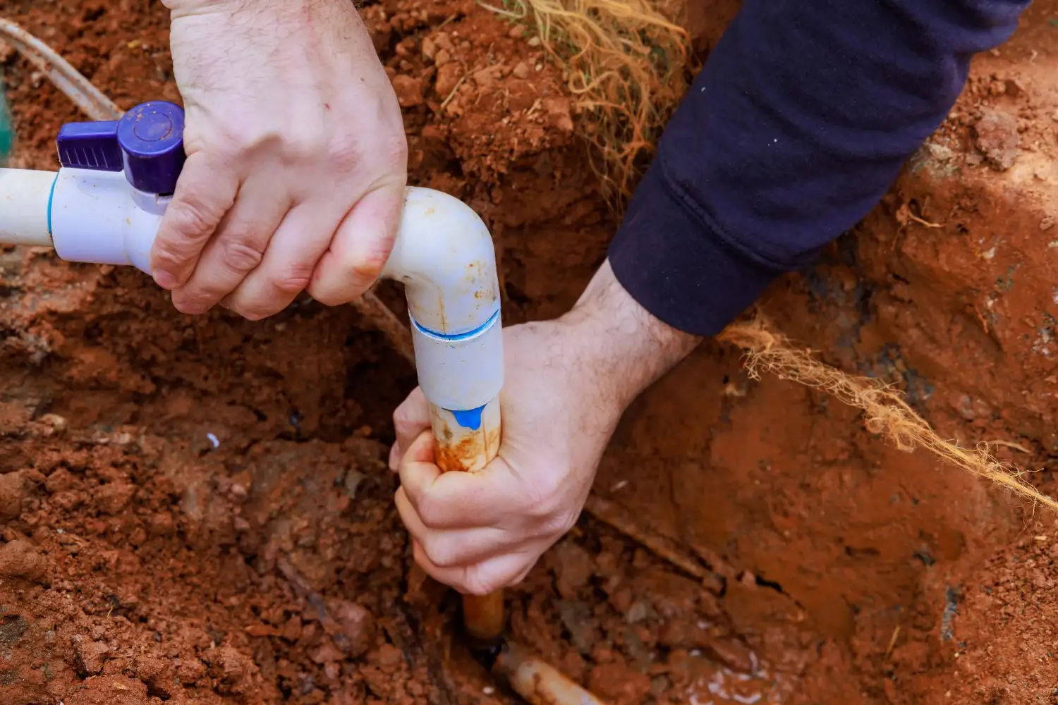 A worker's hands connect a white PVC elbow and a blue valve to a pipe in a narrow trench filled with red-brown soil.