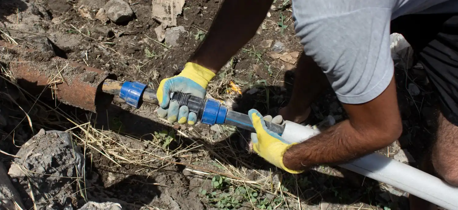 A worker's gloved hands connect a blue and black compression fitting to a rusted metal pipe and a new white pipe in a dirt trench.